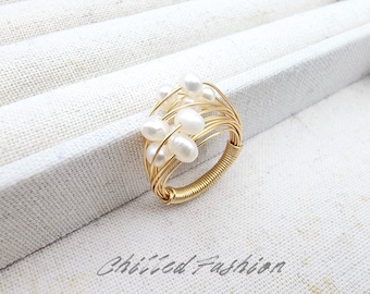 May include: A gold-coloured wire ring adorned with a cluster of white freshwater pearls. The ring's band is tightly wrapped with gold wire, and the pearls are arranged in a delicate, organic design. The ring is set against a white background.