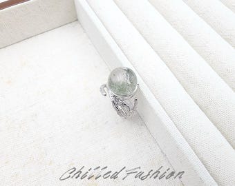 May include: A silver-toned ring featuring a round, clear gemstone with green inclusions. The band has an organic, textured design resembling branches or leaves. The text "Chilled Fashion" is printed in a cursive font at the bottom of the image.