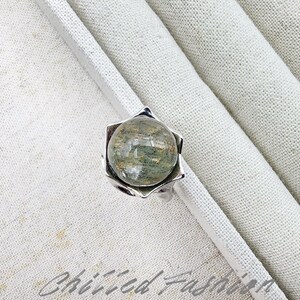 May include: A silver ring featuring a clear, round gemstone with green and brown inclusions. The gemstone is set in a floral-shaped bezel. The ring is displayed on a white, textured surface. The text "Chilled Fashion" is visible.