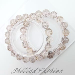 May include: Two bracelets with clear, round beads with a speckled pattern. The bracelets are displayed on a clear plastic holder, resting on a white surface. The beads have a light pink hue.