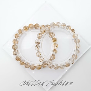 May include: Two clear bracelets with golden-brown beads are displayed on a clear, diamond-shaped surface. The bracelets are circular, and the beads have a speckled appearance. The words "Chilled Fashion" are written in cursive at the bottom.