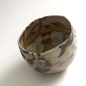 Puede incluir: Un cuenco de cerámica hecho a mano con forma irregular. El exterior presenta una base beige con patrones abstractos en marrón, gris y verde pálido. El interior tiene un esmalte amarillo-verdoso. Ideal para servir o exhibir.