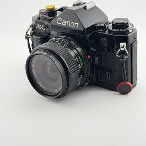 Puede incluir: Una cámara de película Canon A-1 de 35mm en color negro con un objetivo Canon de 35mm f/2.8. El cuerpo de la cámara presenta un botón de disparo dorado, una empuñadura negra texturizada y un pequeño accesorio circular rojo unido a la anilla lateral. El objetivo muestra marcas de texto en blanco y verde.