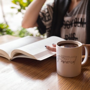 May include: White ceramic coffee mug with black text that reads "#blessed and highly favored". The mug is filled with coffee and sitting on a wooden table.