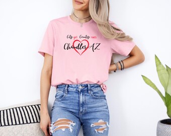 Może przedstawiać: Jasnoróżowa koszulka z napisem "City girl Country roots" nad "Chandler AZ" w czerwonym sercu. Koszulka jest połączona z niebieskimi dżinsami.