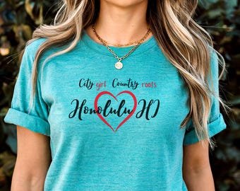 Może przedstawiać: Turkusowa koszulka z podwiniętymi rękawami z napisem "City girl Country roots" nad "Honolulu HI" w czerwonym sercu. Noszony jest złoty naszyjnik.