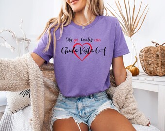 Może przedstawiać: Fioletowa koszulka z napisem "City girl Country roots Chula Vista CA" w kolorze czarnym i czerwonym, z czerwonym sercem. Koszulka jest połączona z dżinsowymi szortami i kremowym kardiganem. Obraz przedstawia osobę.