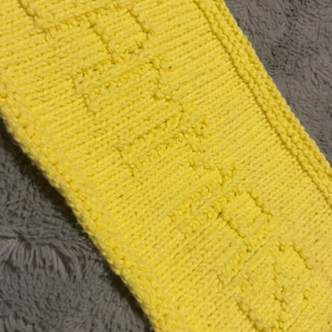 Puede incluir: Un artículo tejido rectangular de color amarillo brillante con la palabra "BABY" tejida en la tela. El artículo tiene una apariencia texturizada y un borde definido. El fondo es un material suave y gris.