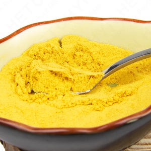 May include: A ceramic bowl filled with yellow powder, possibly a spice or ingredient. A silver spoon rests in the powder, partially scooping it. The bowl has a cream-colored interior with a brown rim and a dark brown exterior.