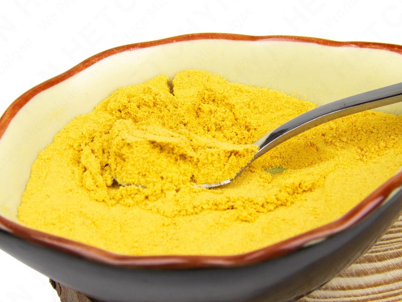 May include: A ceramic bowl filled with yellow powder, possibly a spice or ingredient. A silver spoon rests in the powder, partially scooping it. The bowl has a cream-colored interior with a brown rim and a dark brown exterior.