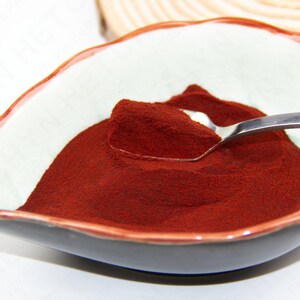 Puede incluir: Un peque&ntilde;o taz&oacute;n blanco con un borde oscuro contiene un mont&oacute;n de polvo rojo oscuro. Una cuchara de plata est&aacute; parcialmente sumergida en el polvo.