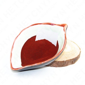 Puede incluir: Un peque&ntilde;o cuenco de cer&aacute;mica blanca con un borde rojo, con forma de hoja, lleno de un polvo rojo oscuro. El cuenco est&aacute; sobre una base de madera.