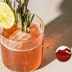 Puede incluir: Un vaso de bebida de color rojo con una rodaja de lima y una ramita de romero como guarnici&oacute;n. La bebida es de un color rojo vibrante y parece ser un c&oacute;ctel o un mocktail. En la esquina inferior derecha se muestra un primer plano del polvo rojo utilizado en la bebida.