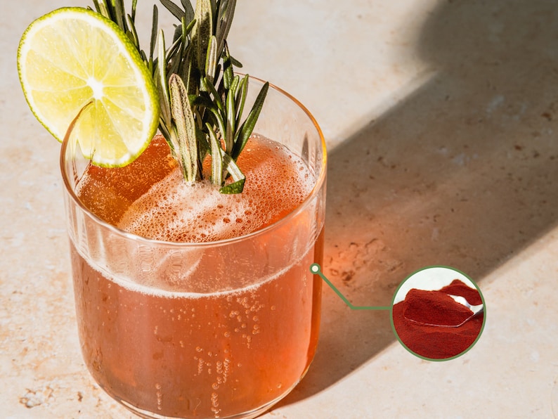 Puede incluir: Un vaso de bebida de color rojo con una rodaja de lima y una ramita de romero como guarnici&oacute;n. La bebida es de un color rojo vibrante y parece ser un c&oacute;ctel o un mocktail. En la esquina inferior derecha se muestra un primer plano del polvo rojo utilizado en la bebida.