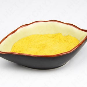 May include: A leaf-shaped ceramic bowl with a black exterior, cream-colored interior, and a reddish-brown rim. The bowl is filled with a bright yellow powder, likely a spice or ingredient.