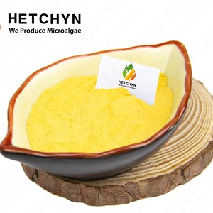 May include: A yellow powder fills a brown and cream-colored bowl, resting on a wooden slice. A small white card with the logo "HETCHYN We Produce Microalgae" is inside the bowl. The bowl has a unique, leaf-like shape.
