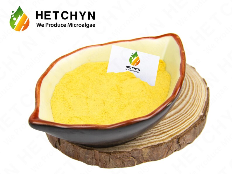 May include: A yellow powder fills a brown and cream-colored bowl, resting on a wooden slice. A small white card with the logo "HETCHYN We Produce Microalgae" is inside the bowl. The bowl has a unique, leaf-like shape.