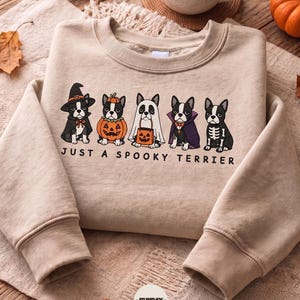 May include: A beige crewneck sweatshirt featuring a graphic of five Boston Terrier dogs in Halloween costumes, including a witch, pumpkin, ghost, vampire, and skeleton. The text below the dogs reads, "JUST A SPOOKY TERRIER."
