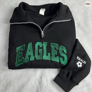May include: A black quarter-zip sweatshirt featuring the word EAGLES embroidered in green across the chest. The right sleeve displays the text DOBALIS above a graphic of a soccer ball. The garment has a silver-toned metal zipper pull.