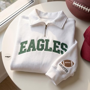 May include: A white quarter-zip sweatshirt featuring the word EAGLES embroidered in dark green block letters across the chest. The sleeve displays an embroidered brown football graphic above the name GRIFFIN in dark green text.