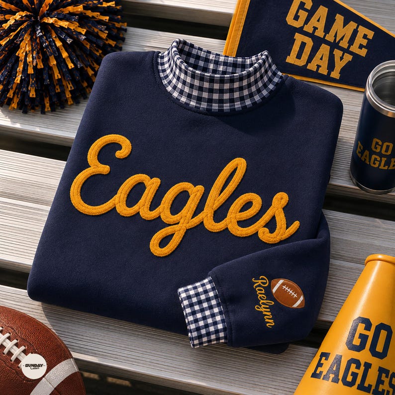 May include: A navy blue sweatshirt featuring the word Eagles in yellow script embroidery across the chest. The garment has a mock neck and sleeve cuffs in a navy and white gingham pattern. The name Raelynn and a football icon are embroidered on the sleeve.