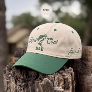 Pode incluir: Boné de beisebol bege e verde com o texto "Reel Cool Dad" e um gráfico de peixe. O boné tem uma aba verde e o nome "Hank" bordado na lateral. O boné está sobre uma superfície de madeira.