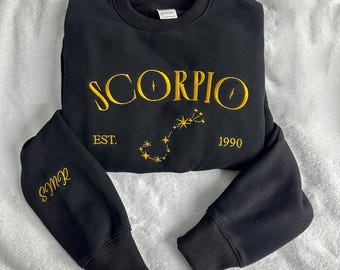 May include: A black crewneck sweatshirt featuring yellow embroidered text that reads SCORPIO, EST. 1990, and SMR on the sleeve. Below the word SCORPIO is a yellow embroidered constellation design with star accents. The garment has long sleeves.