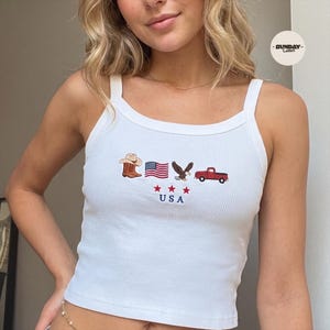 Op de afbeelding: Een wit geribbeld hemdje met een geborduurd ontwerp van een bruine cowboyhoed, een Amerikaanse vlag, een Amerikaanse zeearend en een rode pick-up truck. Onder de iconen staan drie rode sterren en de tekst USA in het blauw.