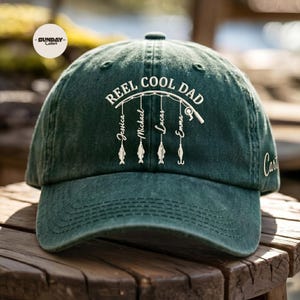 Op de afbeelding: Een donkergroene baseballcap met de tekst "REEL COOL DAD" in het wit geborduurd. Daaronder zijn vissen geborduurd en de namen Jessica, Michael, Lucas en Emma. De pet ligt op een houten oppervlak.