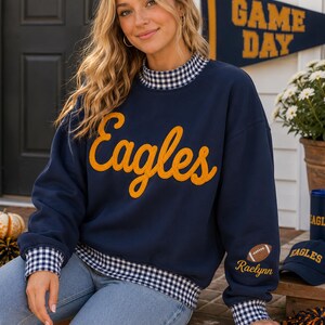 May include: A navy blue long-sleeve sweatshirt featuring the word Eagles in yellow script across the chest. The garment has a blue and white gingham collar, cuffs, and hem. A small football graphic and the name Raelynn are embroidered on the sleeve.