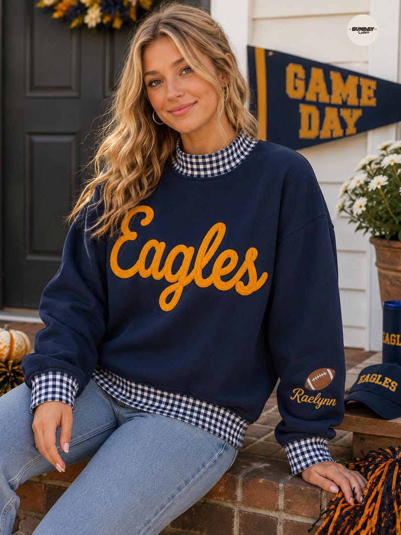 May include: A navy blue long-sleeve sweatshirt featuring the word Eagles in yellow script across the chest. The garment has a blue and white gingham collar, cuffs, and hem. A small football graphic and the name Raelynn are embroidered on the sleeve.