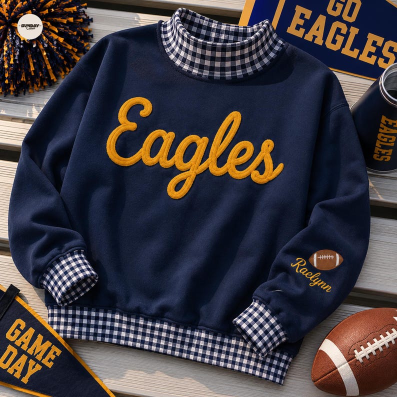 May include: A navy blue long-sleeve sweatshirt featuring a mock neck collar, cuffs, and hem in a navy and white gingham pattern. The word "Eagles" is embroidered in yellow script on the chest, and the name "Raelynn" is embroidered on the sleeve.