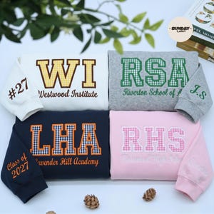 May include: Four sweatshirts in white, gray, navy, and pink, each featuring embroidered varsity-style letters with gingham patterns. The designs include text such as Westwood Institute, Riverton School of A, Lavender Hill Academy, and Rosewood High School.