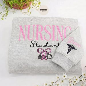 Könnte beinhalten: Ein hellgraues Sweatshirt mit dem Wort "NURSING" in rosa Stickerei über "Student" in Schwarz. Eine rosa Schleife und eine Stethoskop-Grafik befinden sich unter dem Text. Der Ärmel zeigt den Namen "Grace" und ein medizinisches Symbol.