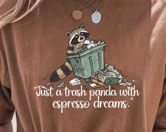 May include: Brown t-shirt featuring a raccoon in a trash can with the text "Just a trash panda with espresso dreams." The raccoon is black, white, and brown. The trash can is green. The shirt is a solid brown color.
