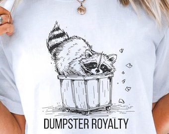 May include: White t-shirt featuring a black and white illustration of a raccoon in a trash can. The text "DUMPSTER ROYALTY" is printed below the graphic. The shirt is a casual, unisex style.