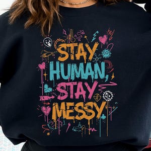 May include: Black sweatshirt featuring the slogan "STAY HUMAN, STAY MESSY" in vibrant, graffiti-style text. The design incorporates hearts, cogs, and other playful illustrations. A relaxed, comfortable garment.