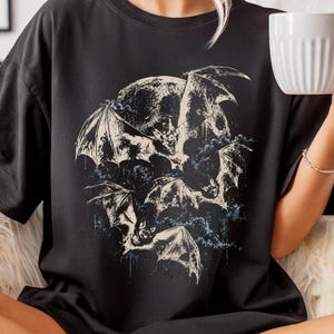 May include: A black short-sleeved t-shirt featuring a detailed graphic print of multiple bats in flight against a large circular moon. The design incorporates dark, textured ink strokes and subtle blue accents, centred on the chest of the garment.