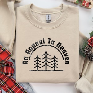 May include: A beige sweatshirt featuring the text "An Appeal To Heaven" in a black circular design with three pine trees. The sweatshirt is folded with a pair of blue jeans and a red plaid bow.