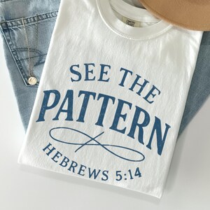 May include: A white t-shirt with the text "SEE THE PATTERN" in blue, along with "HEBREWS 5:14". The shirt is paired with light-wash denim jeans, a tan hat, and a gold necklace. A sprig of dried flowers adds a touch of natural color.