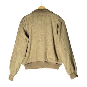 May include: Tan bomber jacket with a ribbed collar, cuffs, and hem. The jacket has a full zipper closure and a textured fabric. The jacket is hanging on a wooden hanger against a white background.