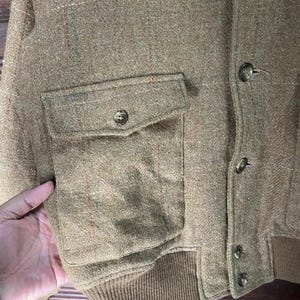 May include: A tan tweed jacket with a button-up front and a single pocket. The jacket has a ribbed hem and cuffs. The fabric has a subtle plaid pattern.