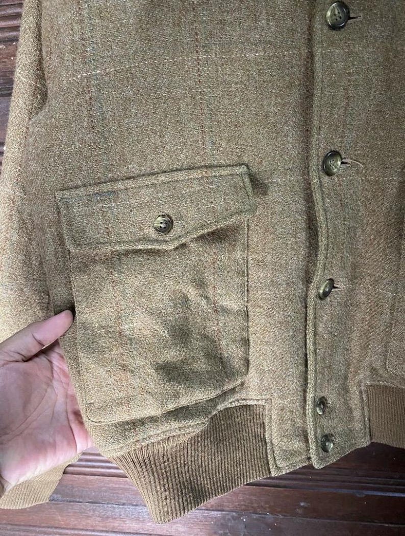 May include: A tan tweed jacket with a button-up front and a single pocket. The jacket has a ribbed hem and cuffs. The fabric has a subtle plaid pattern.