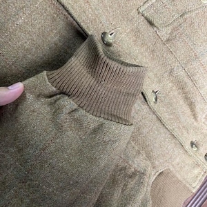 May include: Close-up of a tan jacket with a ribbed cuff and button details. The jacket has a textured fabric and a classic design. The buttons are a matching tan color. The jacket appears to be made of a warm material.