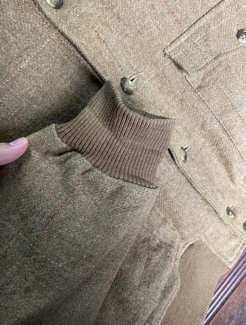 May include: Close-up of a tan jacket with a ribbed cuff and button details. The jacket has a textured fabric and a classic design. The buttons are a matching tan color. The jacket appears to be made of a warm material.