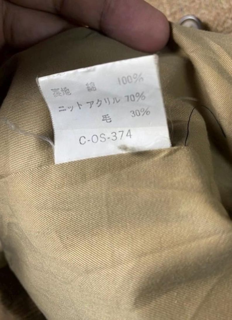 May include: Close-up of a tan fabric with a white tag. The tag has Japanese text indicating the fabric composition: 100% cotton, 70% knit acrylic, and 30% wool. The tag also includes the code C-OS-374.