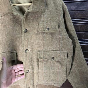 May include: A tan Polo by Ralph Lauren jacket with a button-up front, two chest pockets, and two lower pockets. The jacket has a ribbed collar and cuffs. The brand name is visible on the inside of the jacket.
