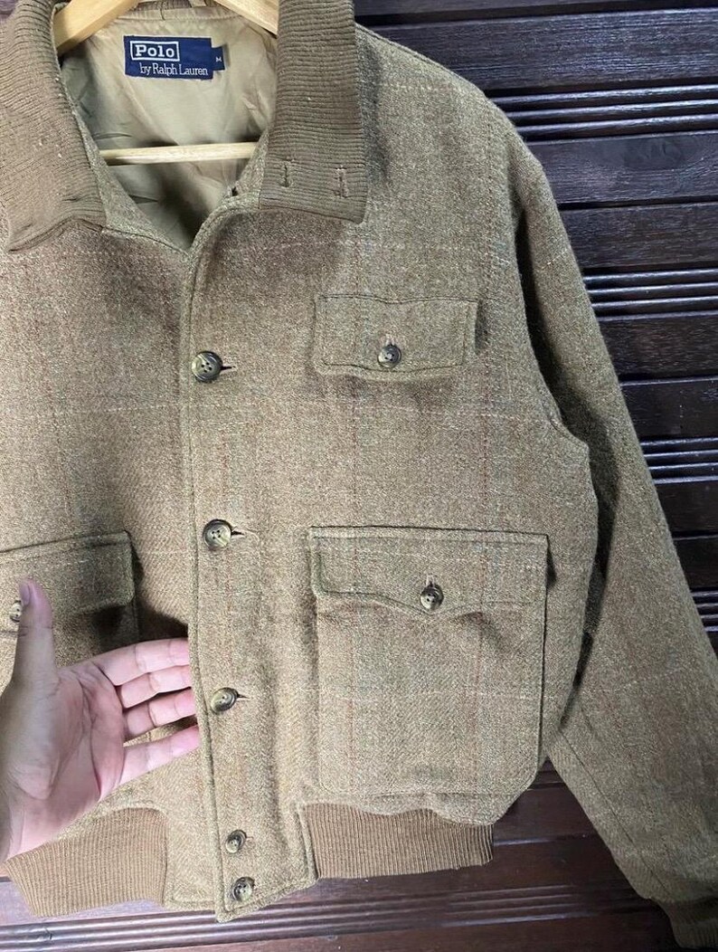 May include: A tan Polo by Ralph Lauren jacket with a button-up front, two chest pockets, and two lower pockets. The jacket has a ribbed collar and cuffs. The brand name is visible on the inside of the jacket.