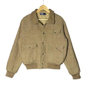 May include: A tan bomber jacket with a brown collar and cuffs. The jacket has two front pockets and a button-up closure. The jacket is hanging on a wooden hanger.