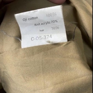 May include: Close-up of a tag with text. The tag reads "Oji cotton 100%, Knit acrylic 70%, hair 30%" and "C-OS-374". The tag is inside a tan fabric.